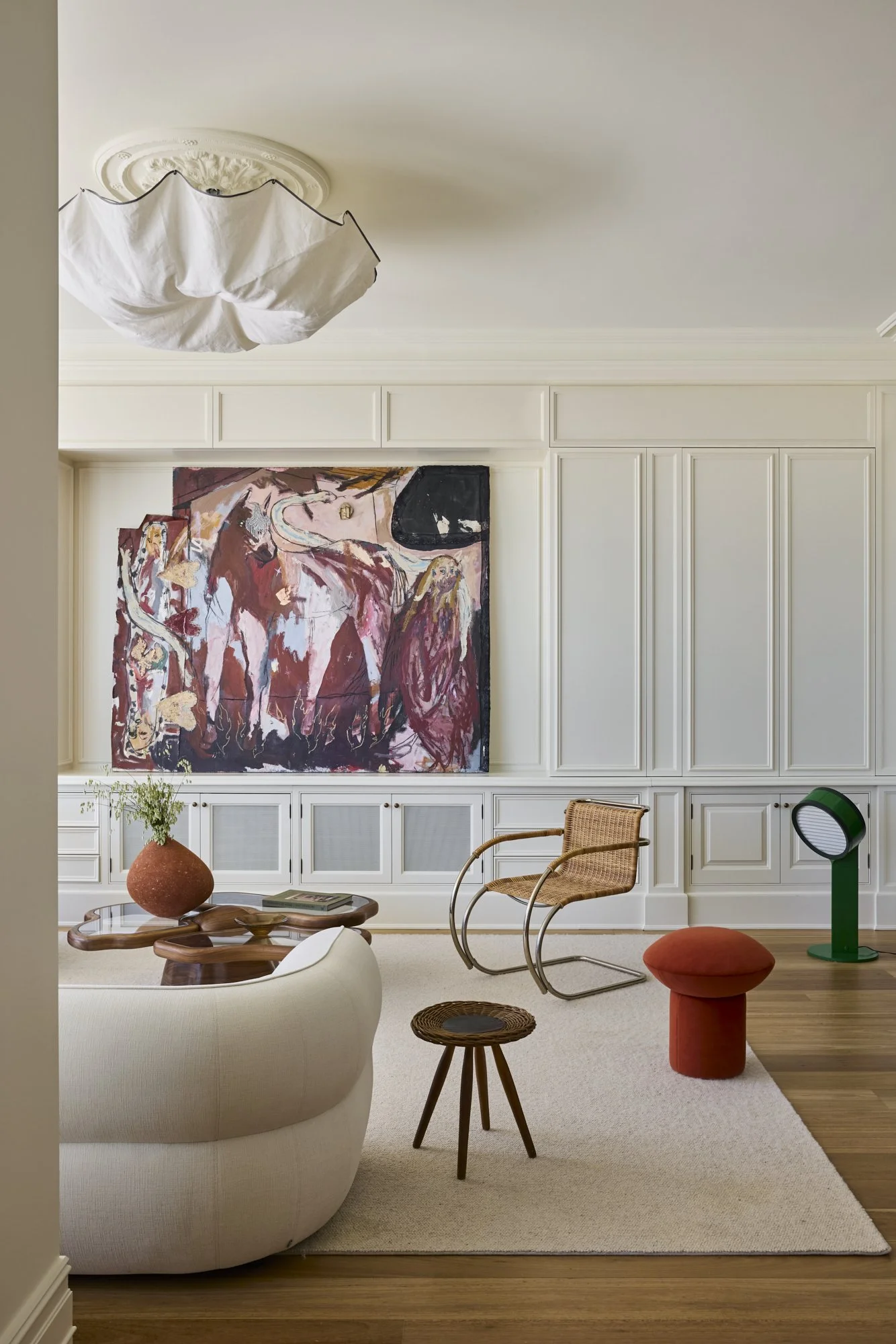 Modern living room with white paneled walls, large abstract painting, white sofa, rattan chair, small wooden stool, green lamp, and orange ottoman on beige carpet, hardwood floor, ceiling light fixture.