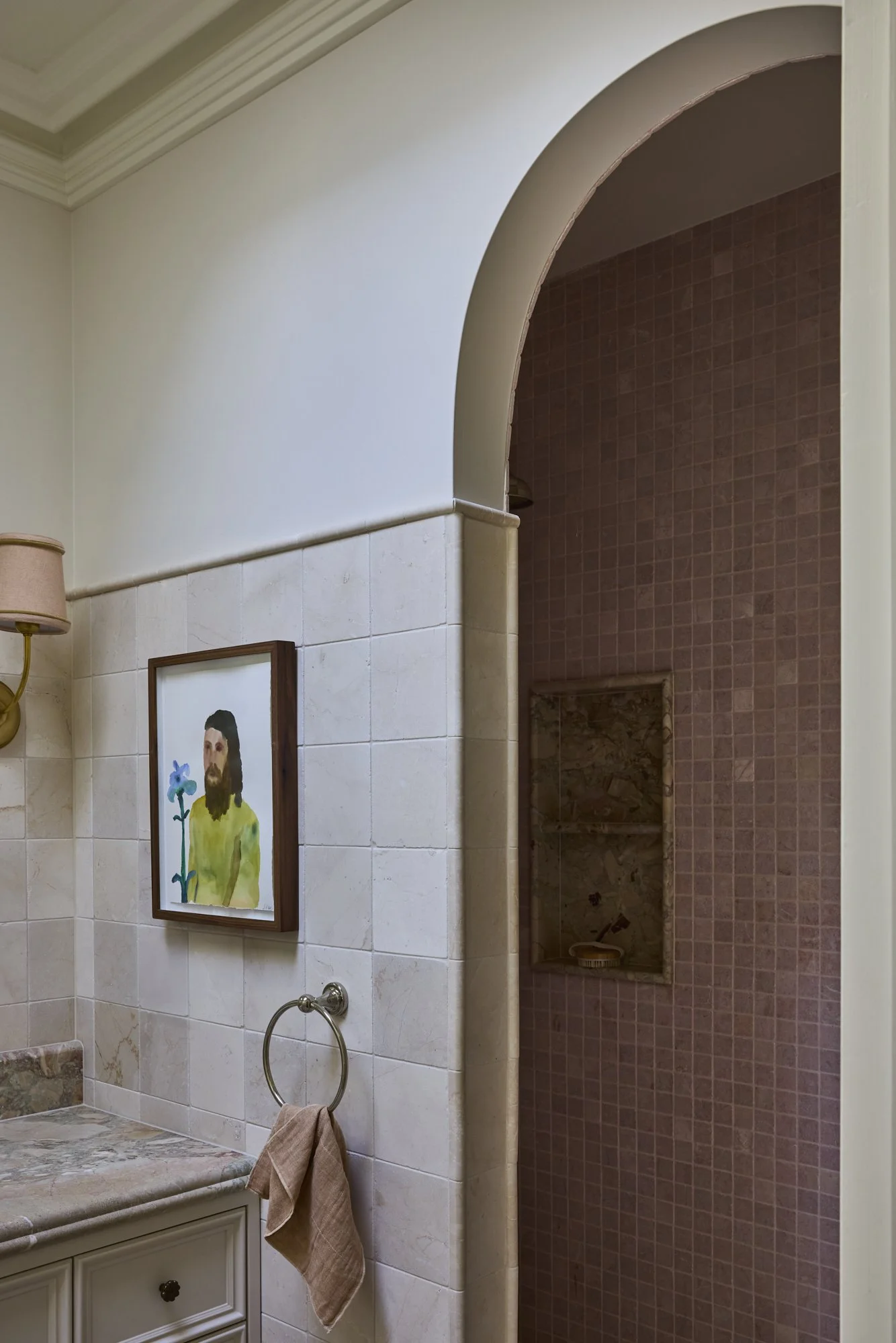 Bathroom with a beige sink and countertop, a framed picture of Jesus with a flower, a wall-mounted lamp with a pink shade, and a towel hanging on a ring. An arched doorway leads to a shower with pink tiles and a built-in niche.