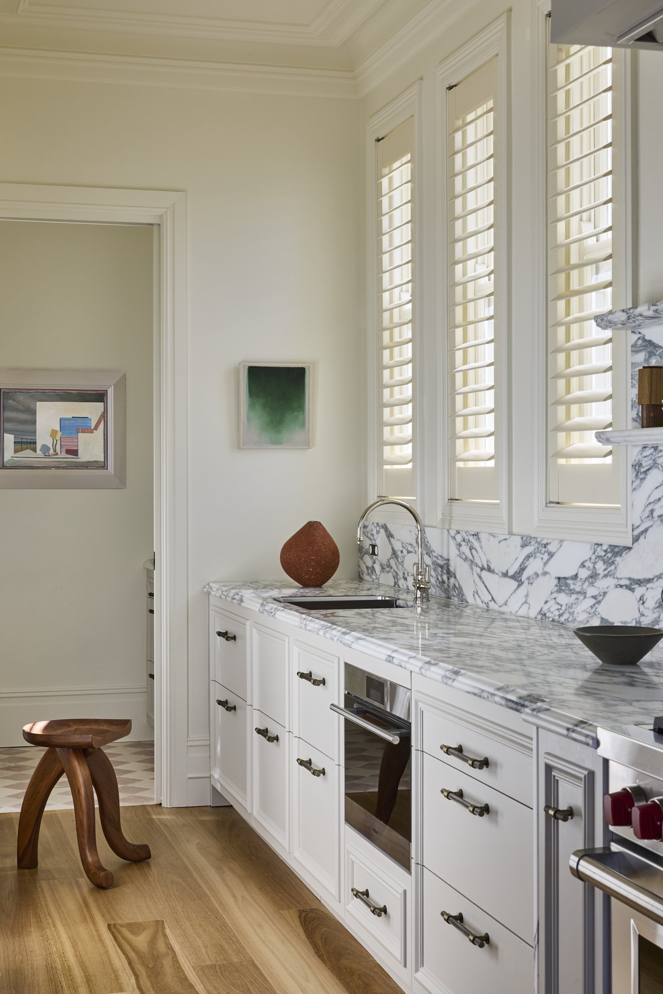 A modern kitchen with white cabinets, marble countertops, and a marble backsplash. There are three windows with white shutters, and a wooden stool in the foreground. Artwork is hanging on the wall.