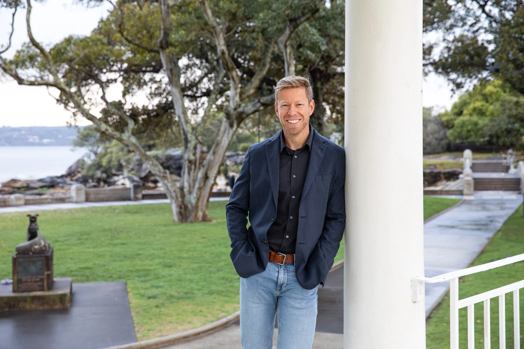 Professional portrait of a smiling man wearing a navy blazer and black shirt, photographed outdoors in a Sydney park setting.