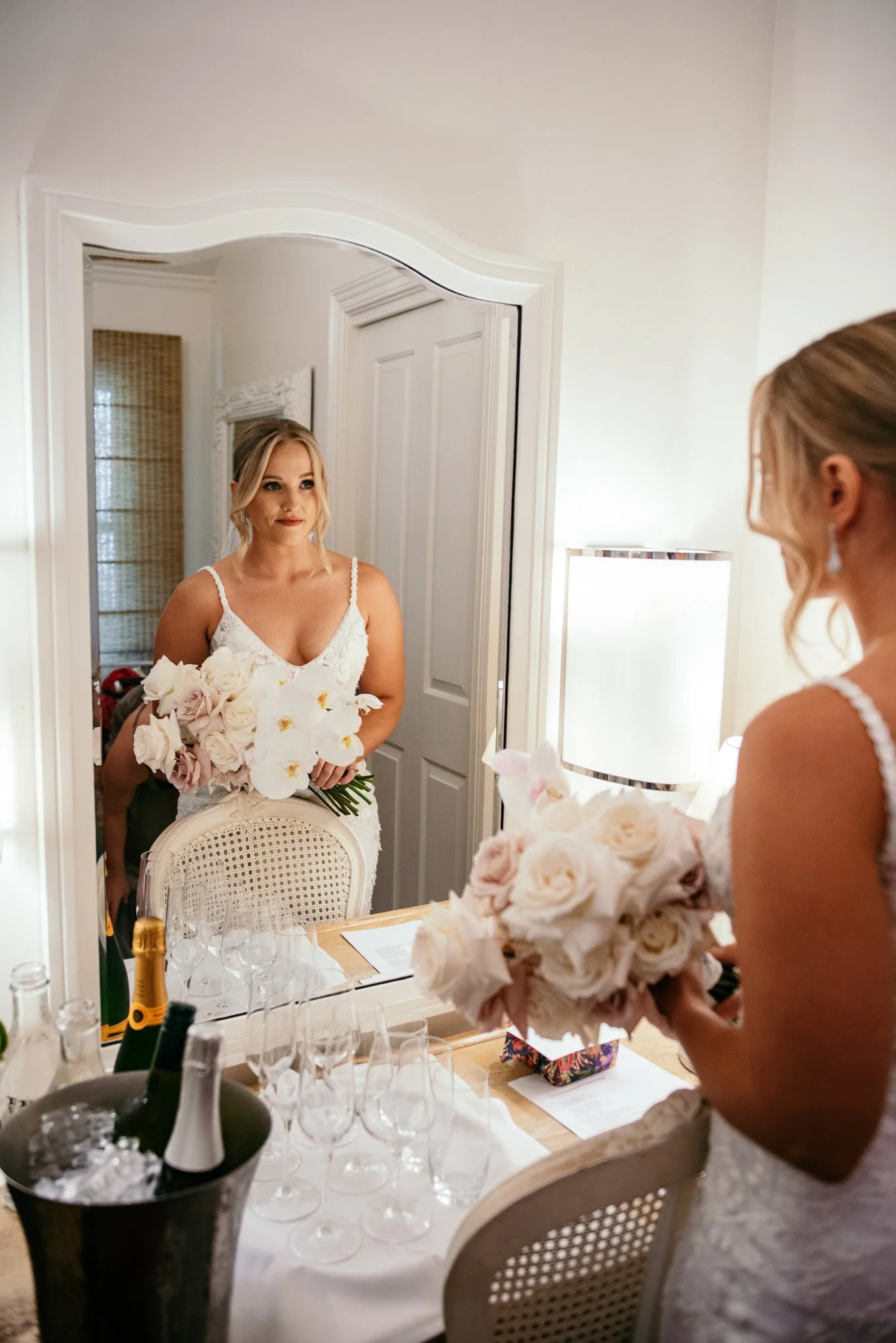 Bride in a white lace dress holding a bouquet of flowers, looking at her reflection in a mirror while standing in a softly lit room with champagne glasses on a table.