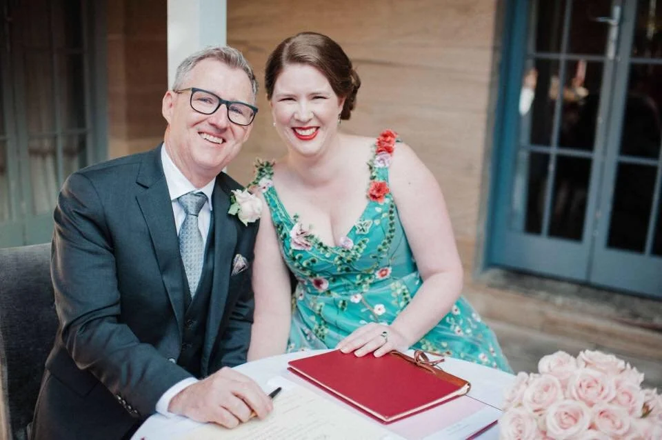 A smiling man in a dark suit and glasses sitting next to a woman in a floral dress, both at a table with a document and pink roses, inside a room with wooden walls and glass doors.