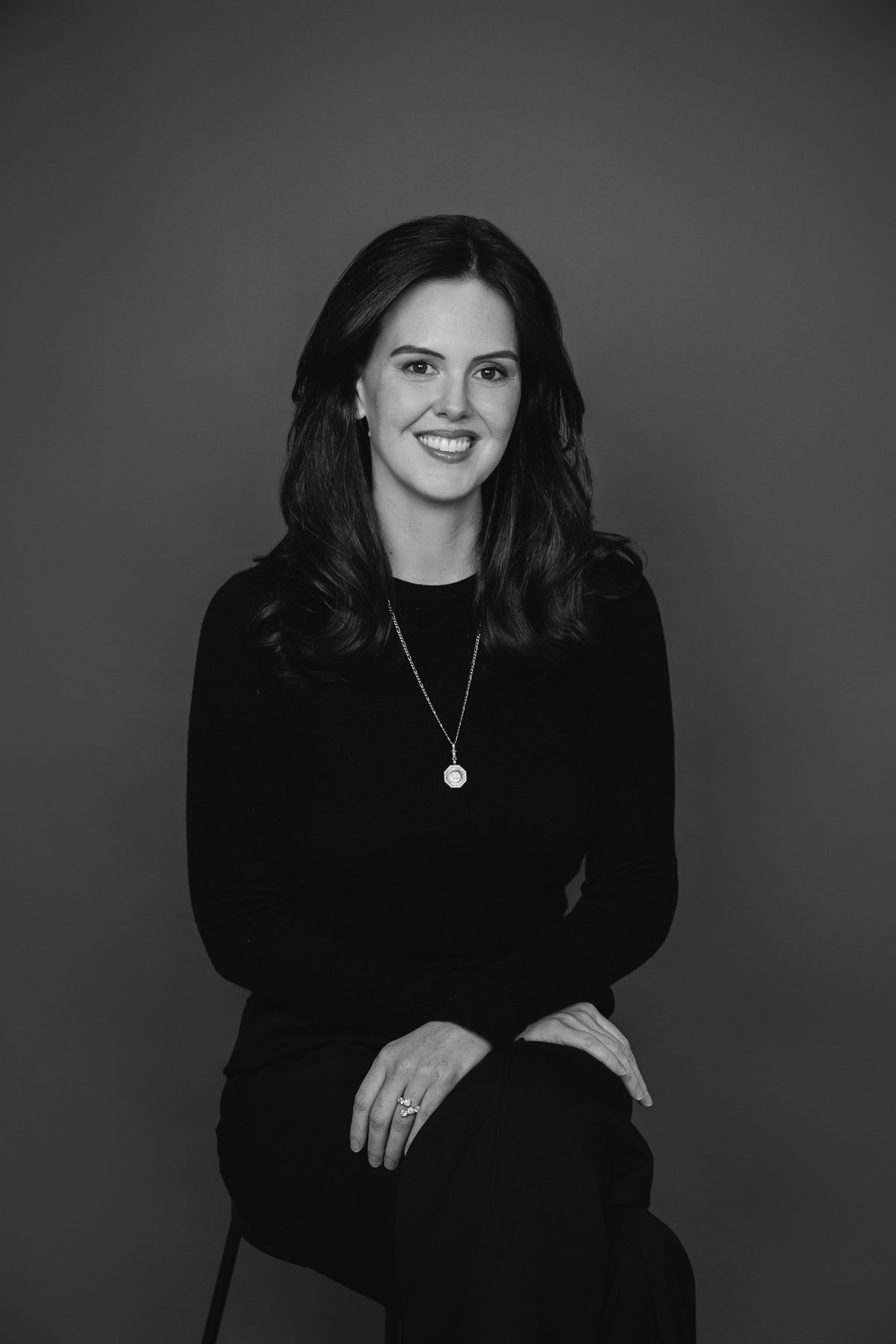 Black and white professional headshot of a woman wearing a simple black outfit and necklace, photographed against a dark studio background.