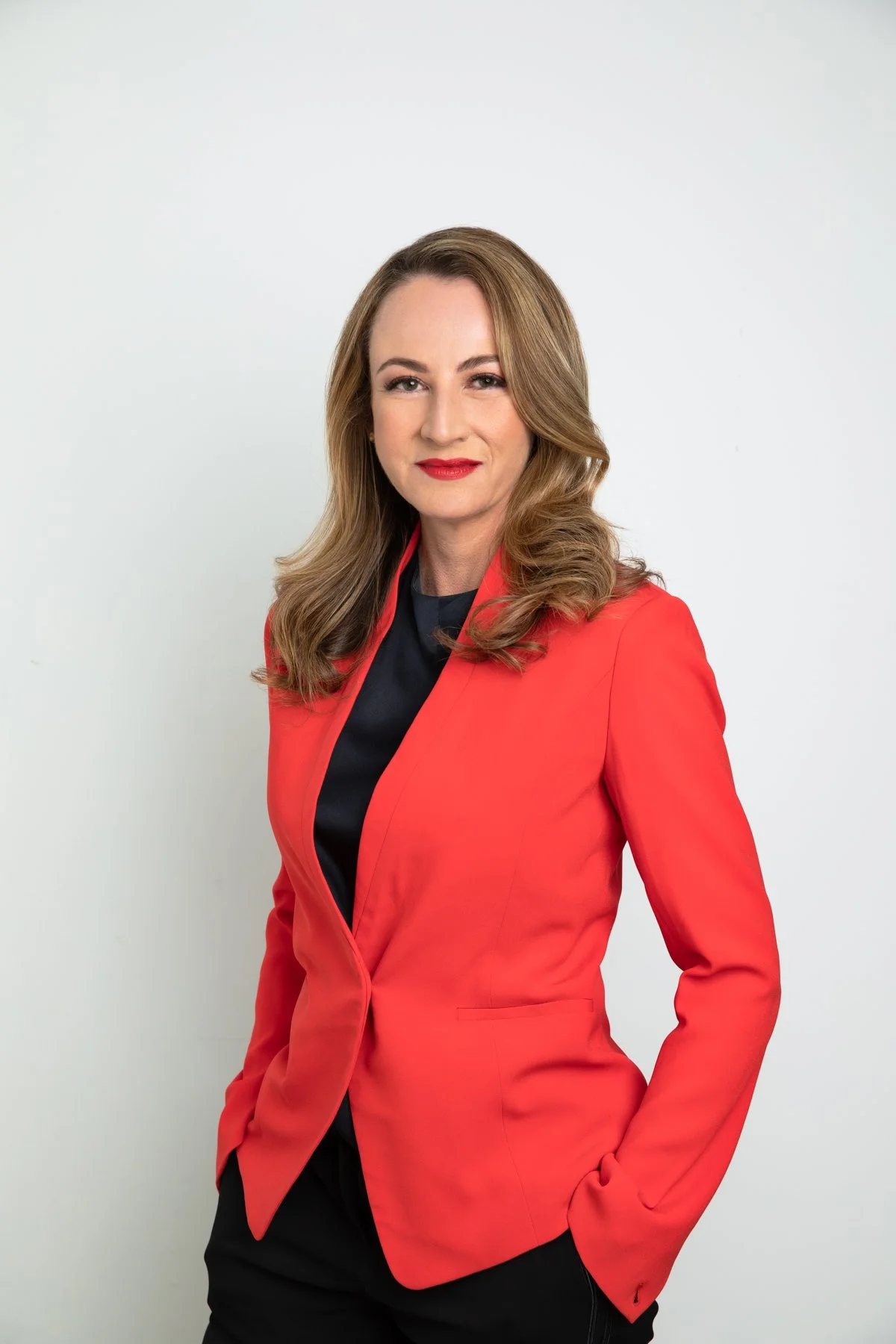 A confident woman with wavy, shoulder-length light brown hair, wearing a red blazer over a black top, standing against a plain white background.