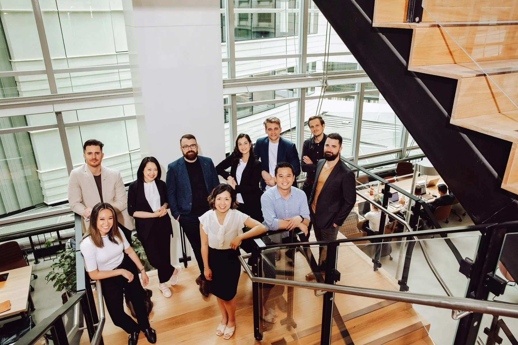 A diverse group of ten professionals posing on a staircase inside a modern office building with large glass windows, wooden floors, and open workspace area in the background.