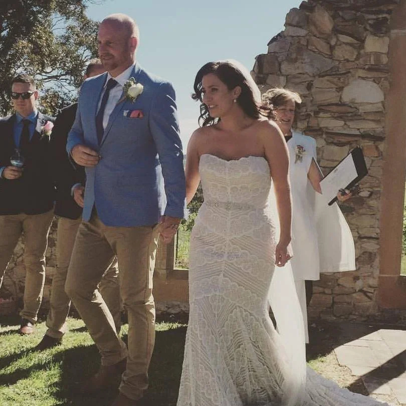 A bride and groom walking together during a wedding ceremony outdoors, with a small group of guests and a stone wall in the background.