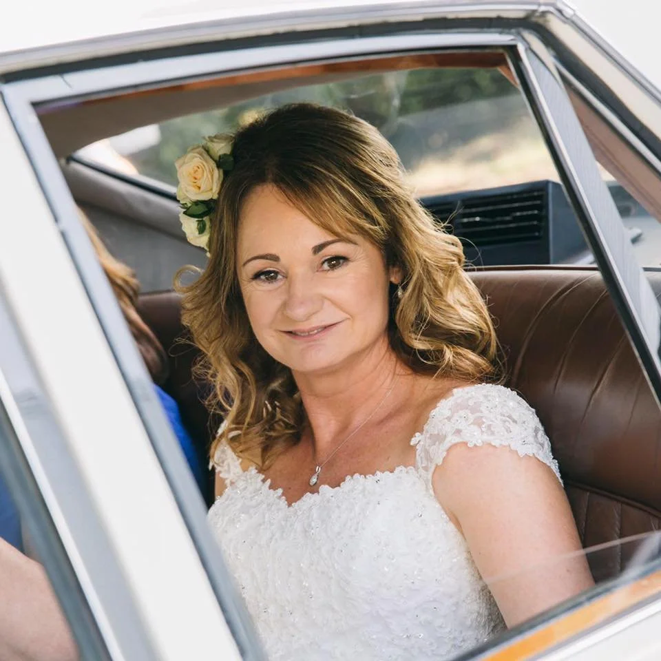 A woman in a white wedding dress sitting in the back of a vintage car with a flower in her hair.