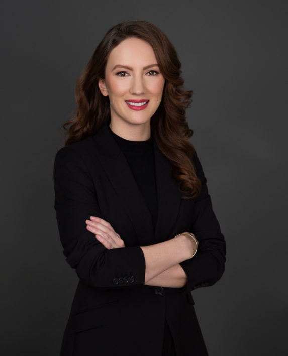 Brunette woman in black professional outfit, smiling against gray background.
