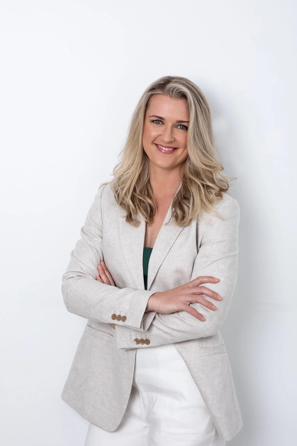 Professional headshot of a woman in a beige blazer with arms crossed, smiling against a white background