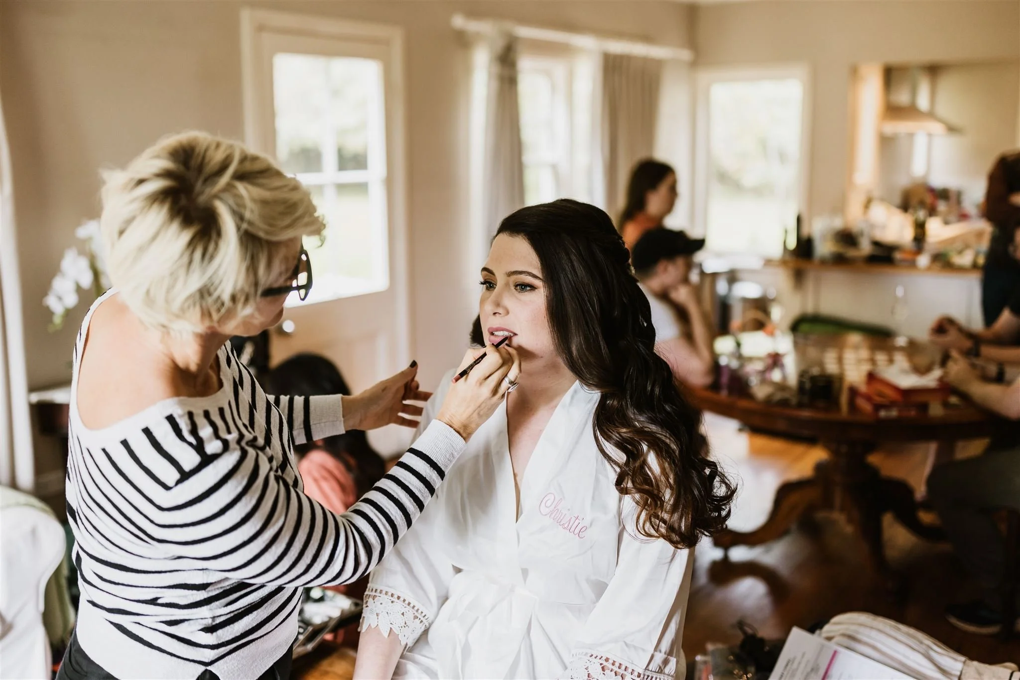 Simona Janek applying bridal makeup on a bride in a white robe during wedding morning preparations
