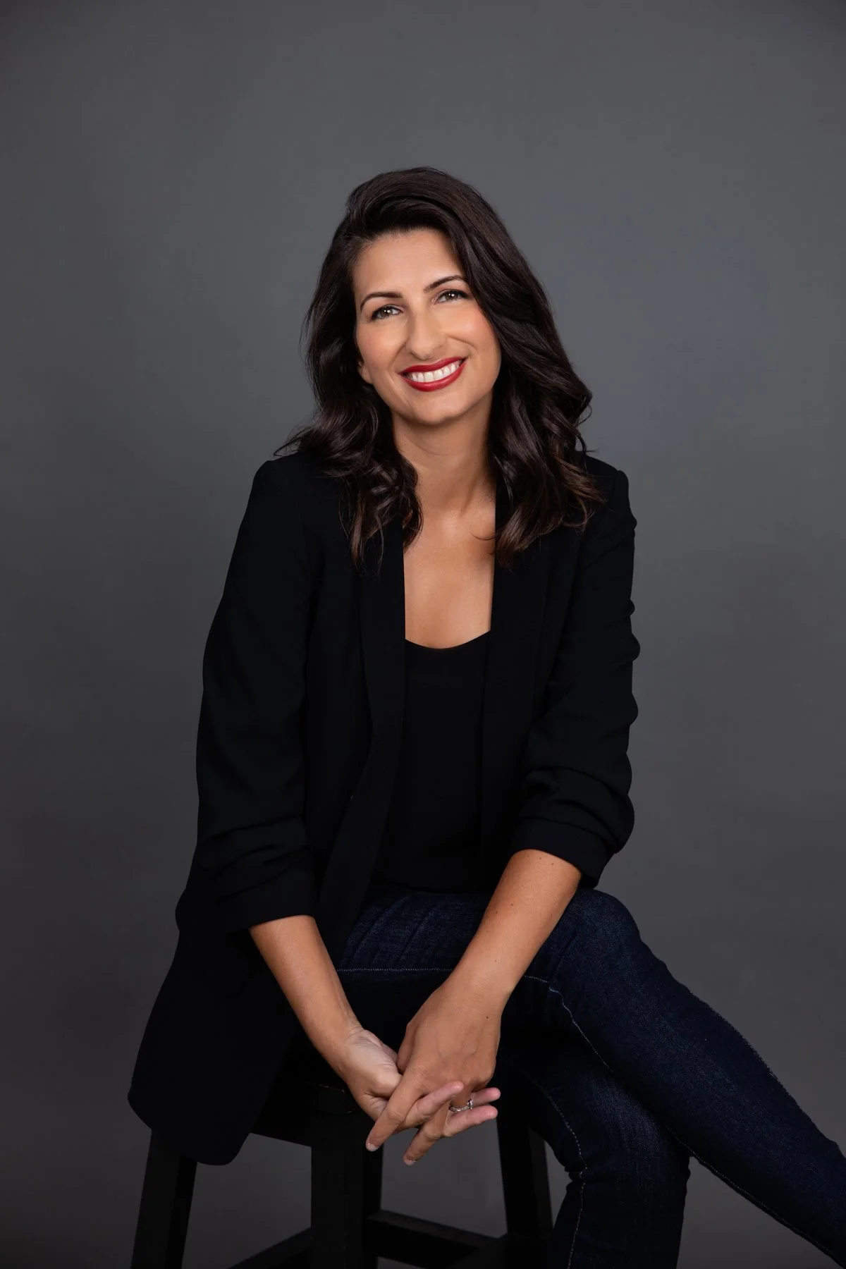 Professional headshot of a woman in a black blazer seated on a stool, smiling confidently against a dark grey background