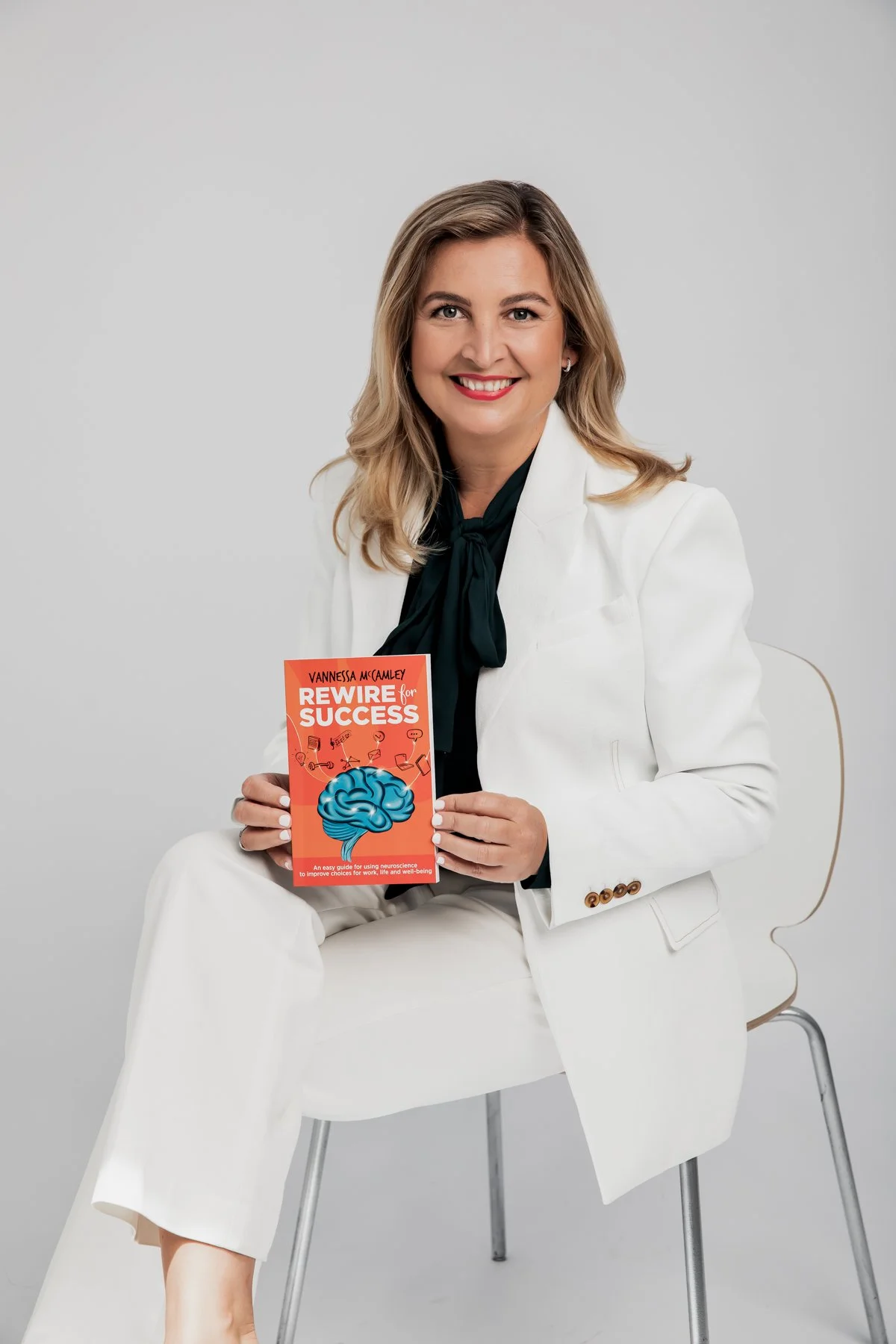 Professional headshot of a smiling woman wearing a white suit and black blouse, holding a book in a Sydney photography studio.