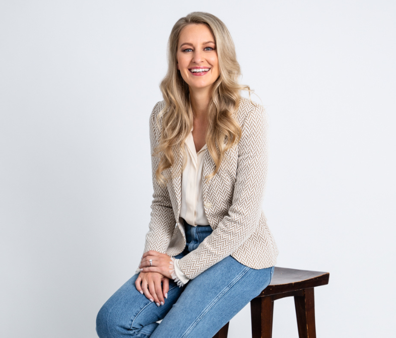 Professional brand photo of a smiling woman in a blazer and jeans seated on a stool against a white background