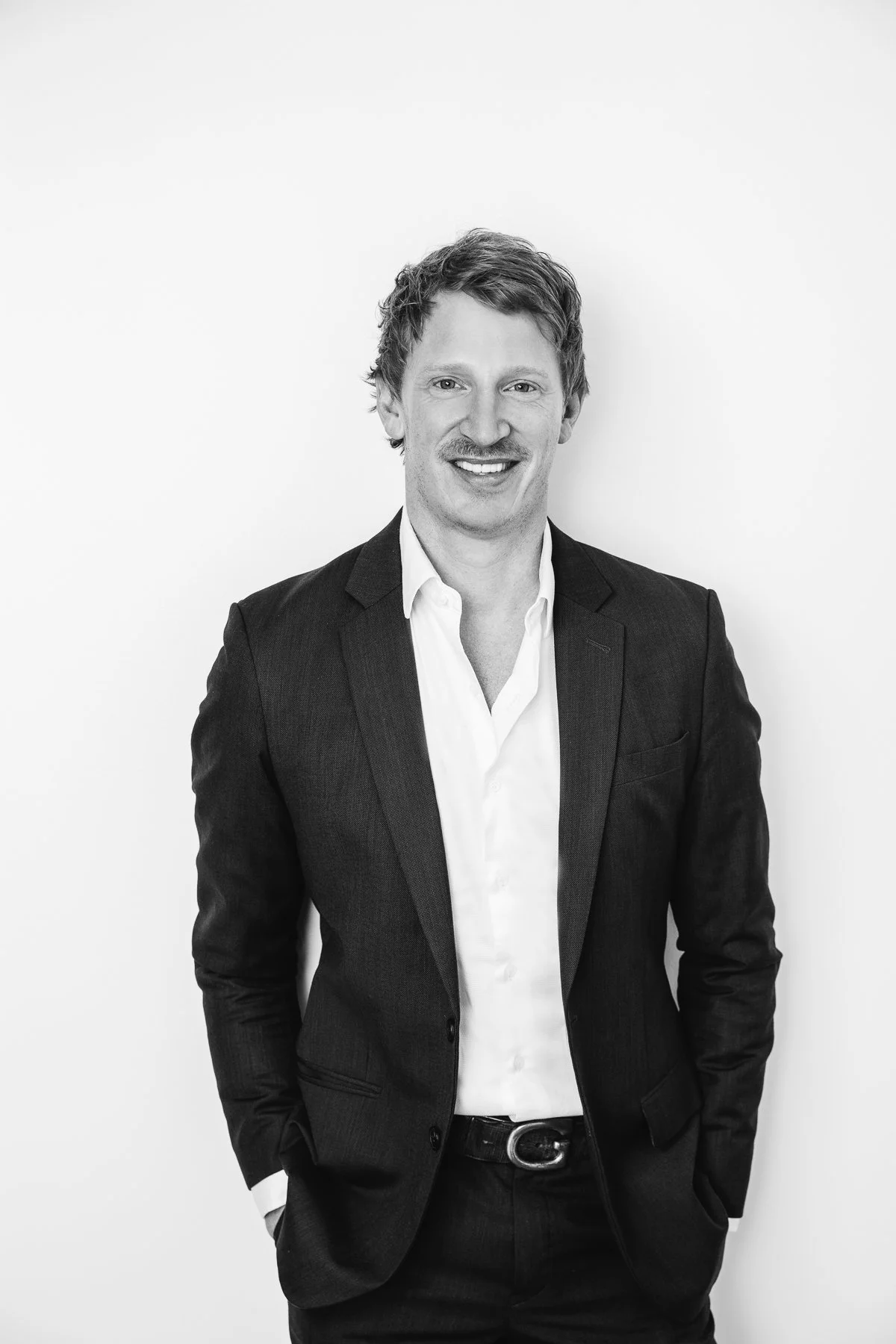 Black and white professional headshot of a man in a dark blazer and open-collar shirt against a white background