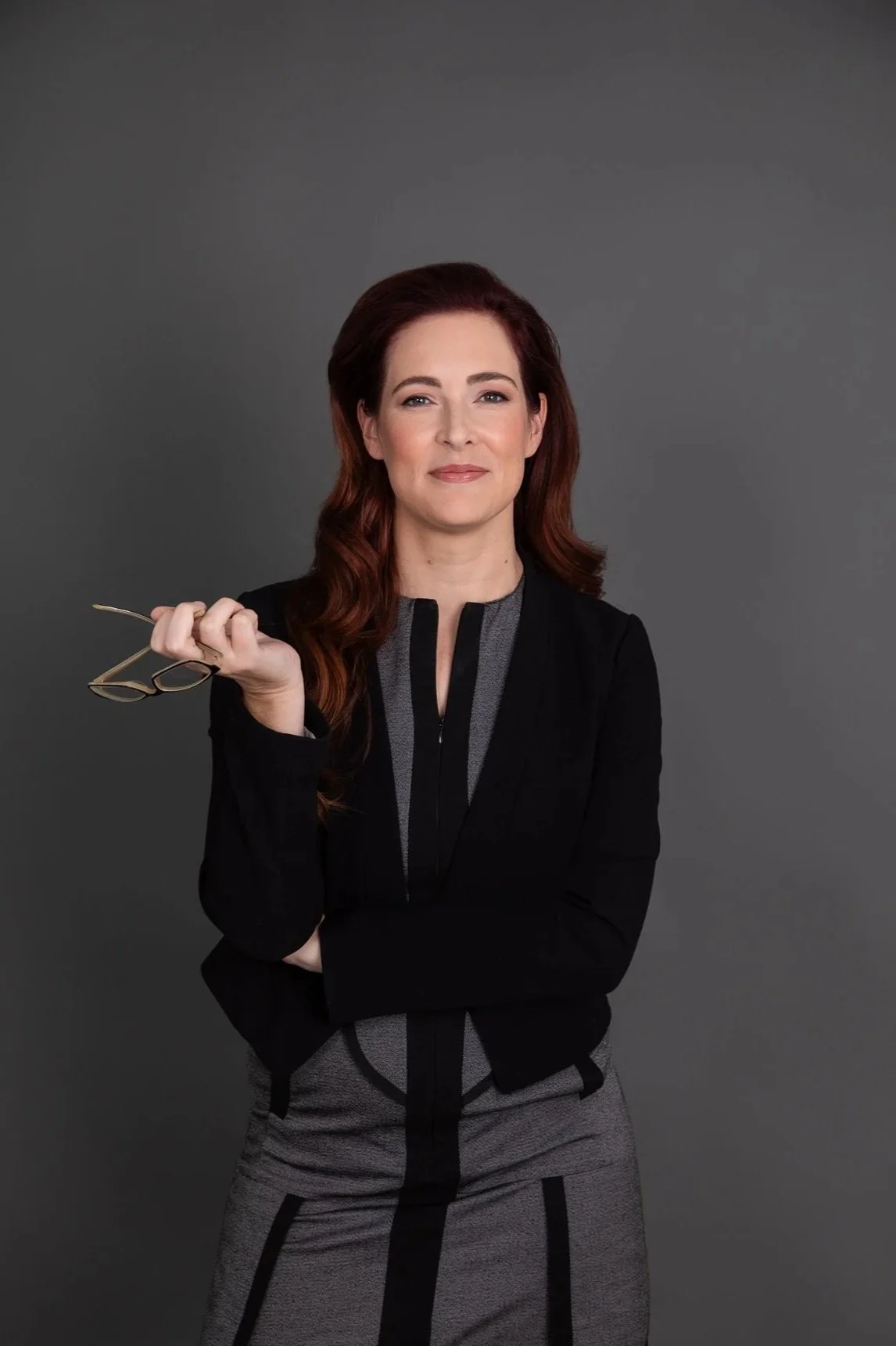 Portrait of a woman with red hair holding glasses, standing against a dark gray background.