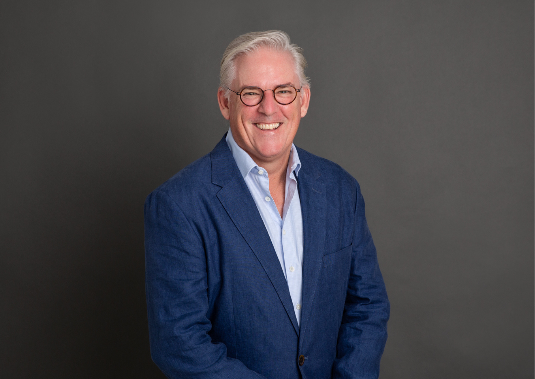 Natural LinkedIn headshot of a smiling professional man wearing a blue blazer and glasses, photographed against a dark studio background in Sydney.
