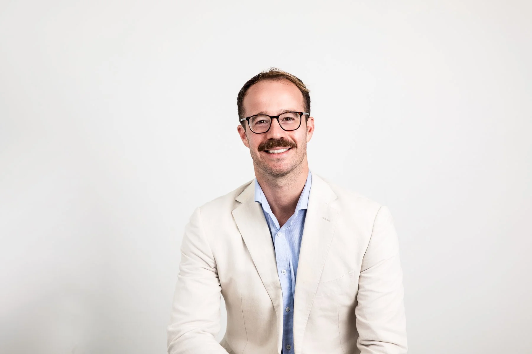 Professional LinkedIn headshot of a smiling man wearing a white blazer and glasses, photographed against a clean light studio background in Sydney.