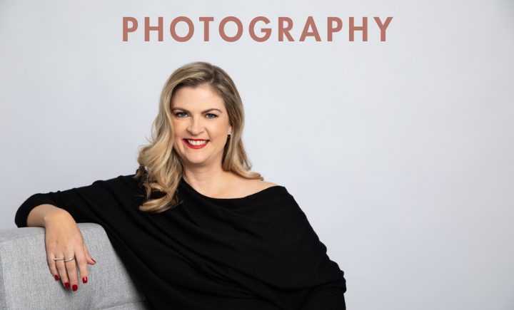 Smiling woman with blonde hair wearing a black off-shoulder top, seated on a light grey chair against a neutral background with the word “PHOTOGRAPHY” above her.