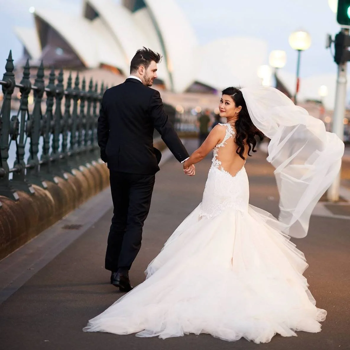 A bride and groom holding hands and looking at each other on a city street at dusk, with the Sydney Opera House in the background.