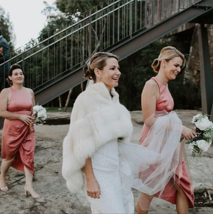 A bride and her bridesmaids walking outdoors near a staircase, dressed in pink and holding bouquets, with trees in the background.