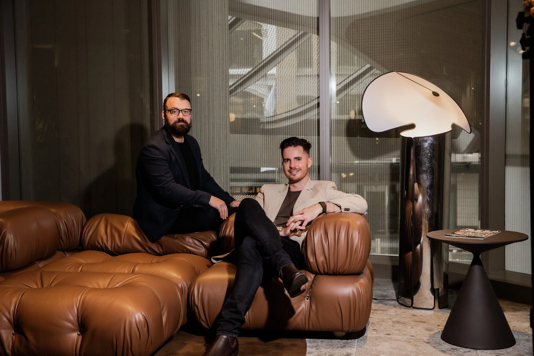 Two men posing confidently on a brown leather sofa in a modern studio setting — professional corporate headshots photography by Simona Janek