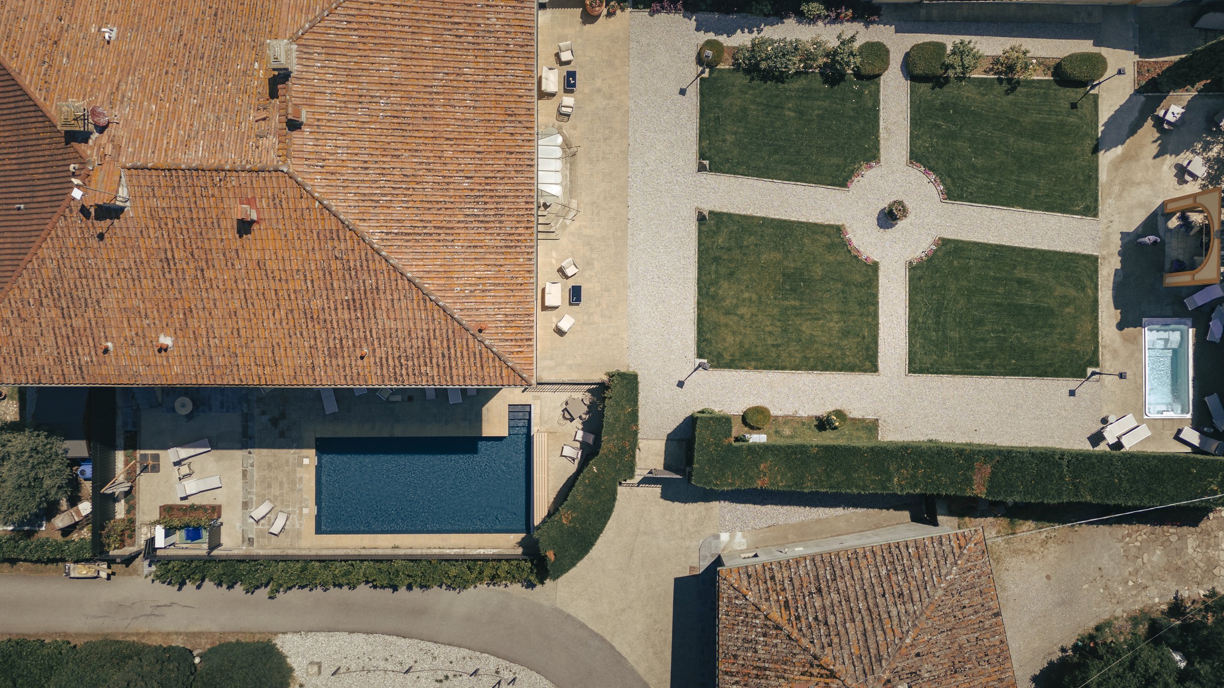 Vista dall'alto di un cortile con prato diviso in quattro parti, con un vialetto che attraversa il centro, panchine e una piscina piccola, con alcuni arredi intorno.