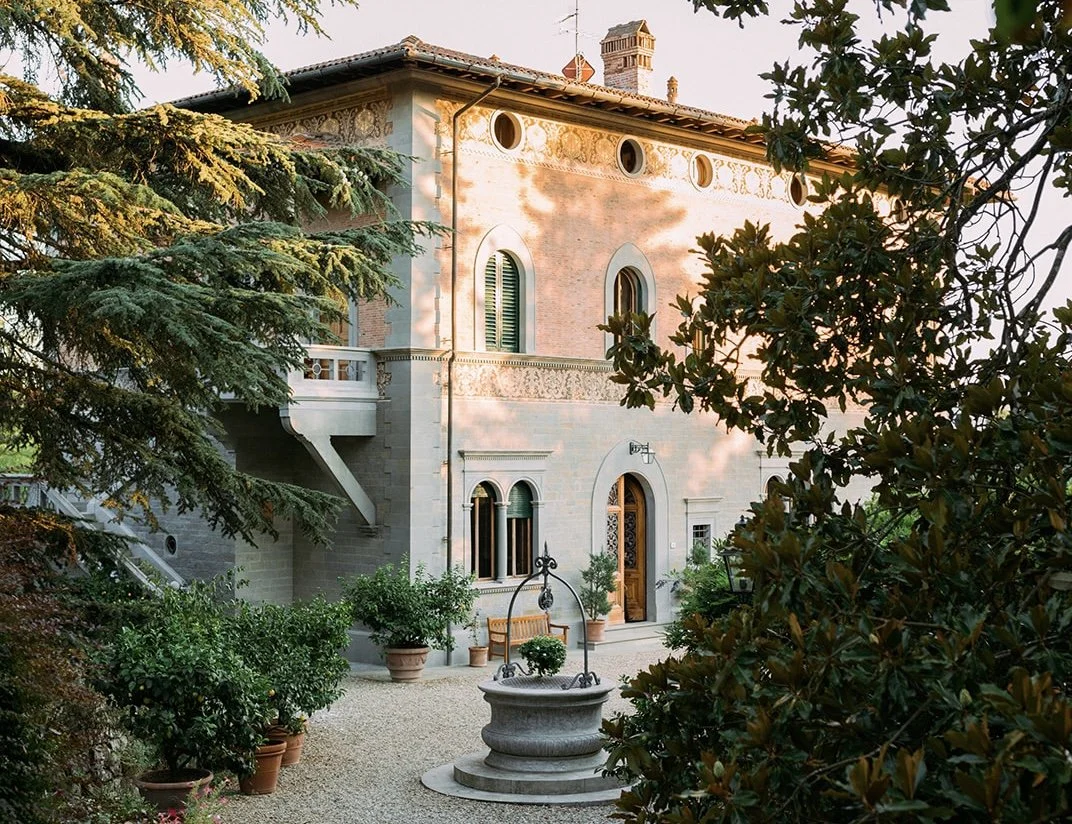 Una villa in stile classico con facciata in pietra, finestre ad arco e una decorazione sottile lungo il tetto. Nel giardino ci sono piante verdi, una fontana in pietra e una panchina di legno circondata da vasi di fiori. Alberi e cespugli incornician