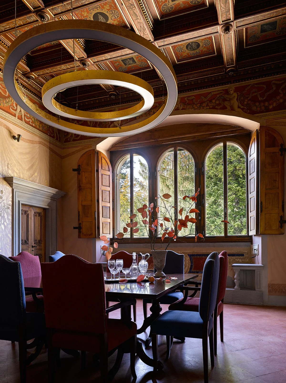 Sala da pranzo con tavolo, sedie di diversi stili, vasi e bicchieri, grande finestra con persiane di legno, decorazioni sul soffitto e pareti.