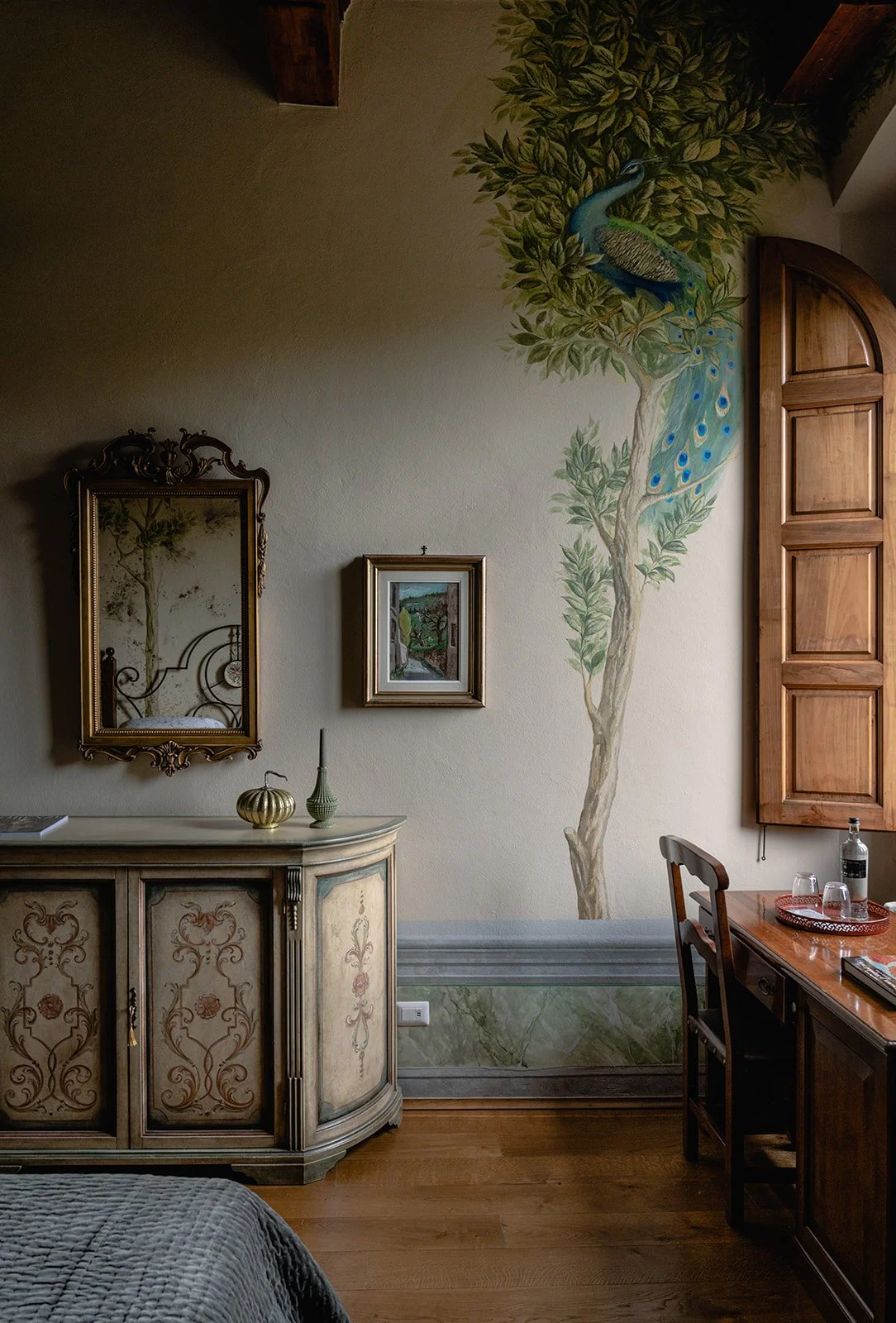 Interno di una stanza con parete decorata con un dipinto di un pavone su un albero. Mobili in legno, specchio dorato sopra un mobile dipinto, piccola decorazione, e parte di un letto visibile in basso a sinistra.