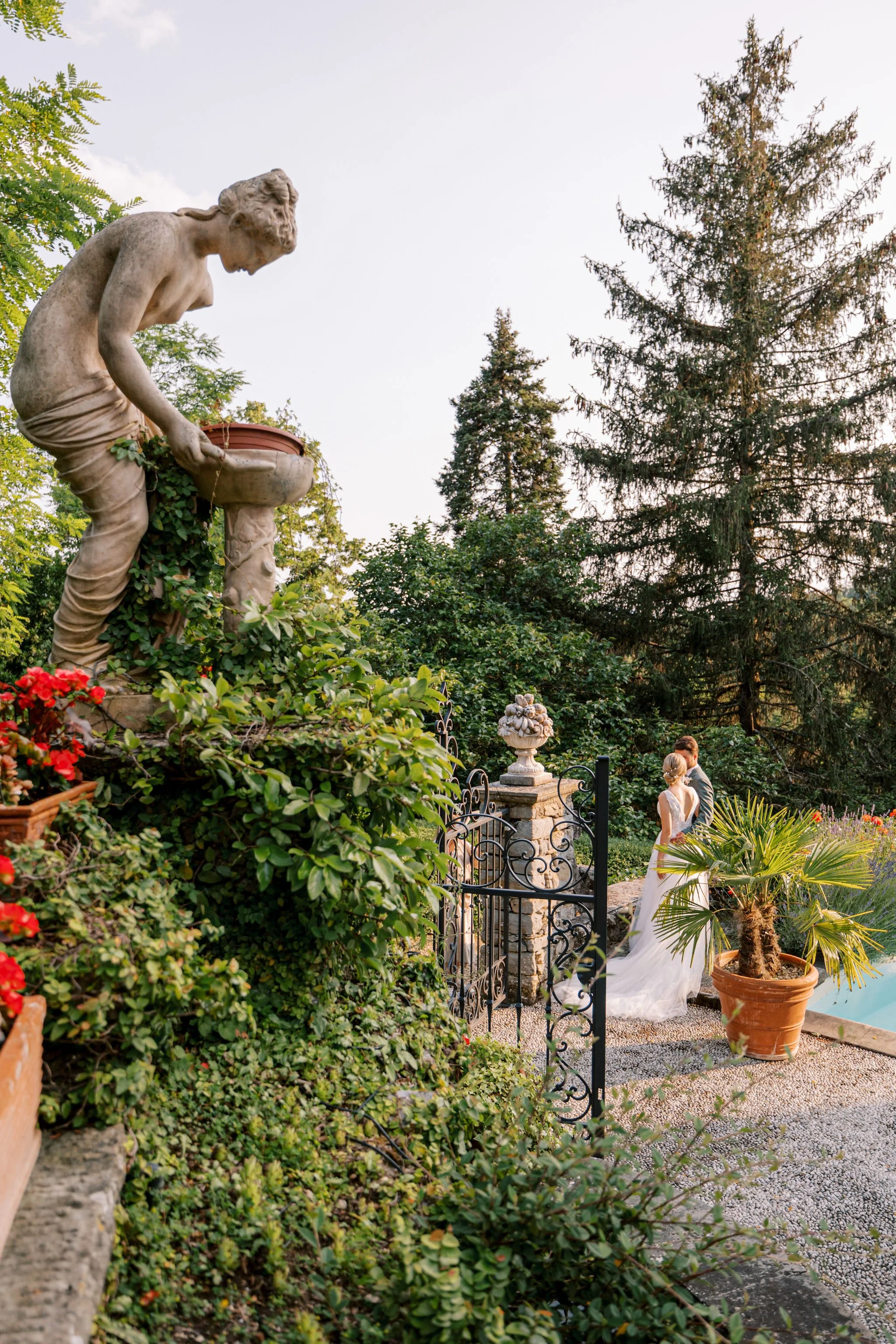 Una coppia di sposi si bacia in un giardino con piante, statue e un cancello in ferro, al tramonto.