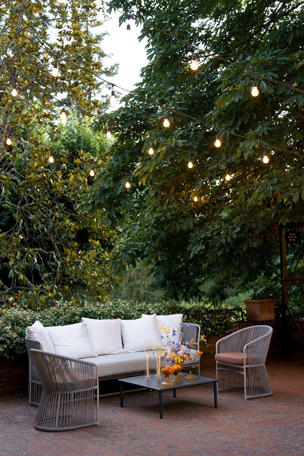 Area all'aperto con divano, tavolino, sedie e decorazioni floreali sotto luci pendenti e alberi verdi.