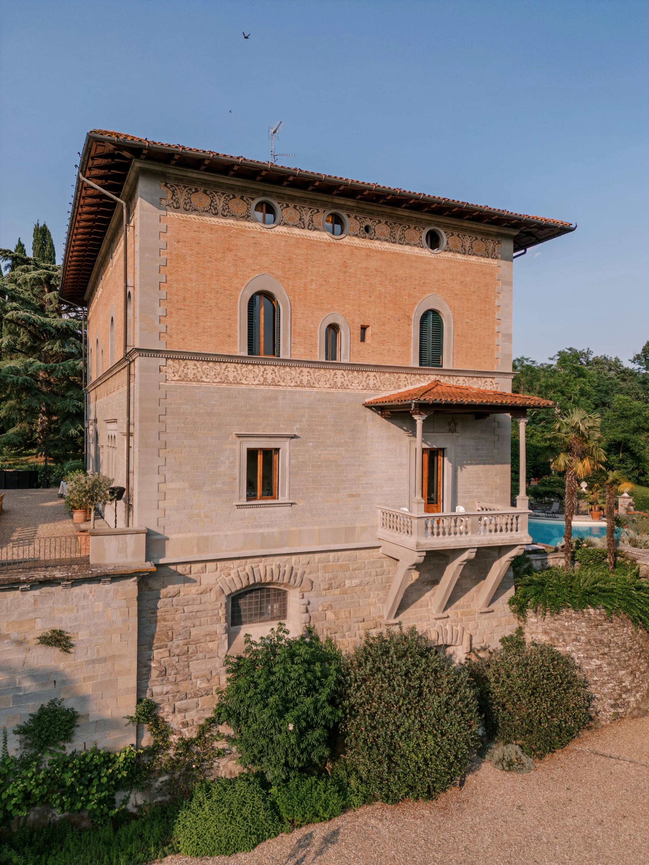 Una villa moderna in stile classico con pareti di mattoni e pietra, balcone con ringhiera, e un giardino con piante verdi e un'area con piscina al lato.