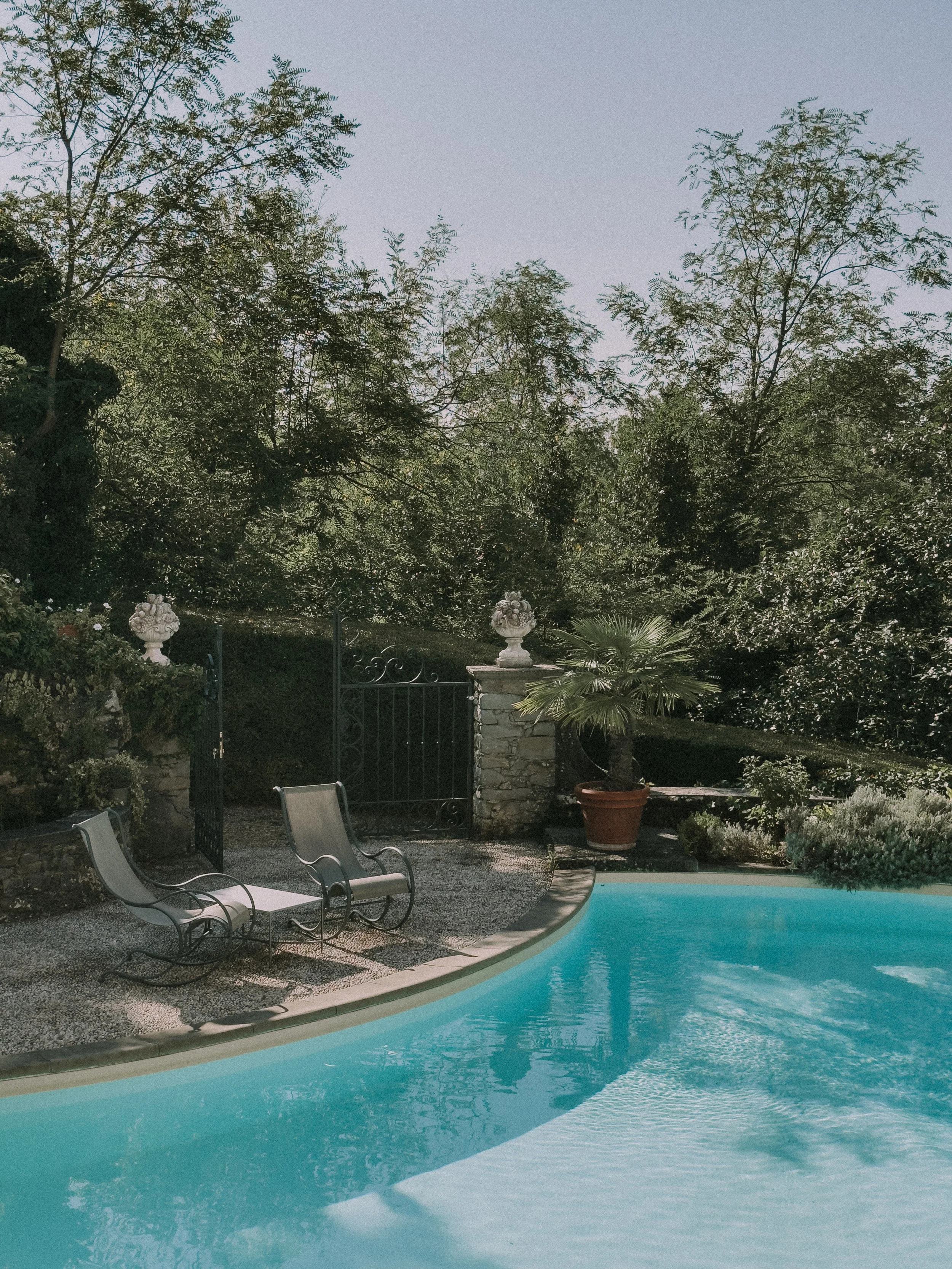 Piscina all'aperto con due lettini e piante verdi, alberi sullo sfondo e cielo chiaro.
