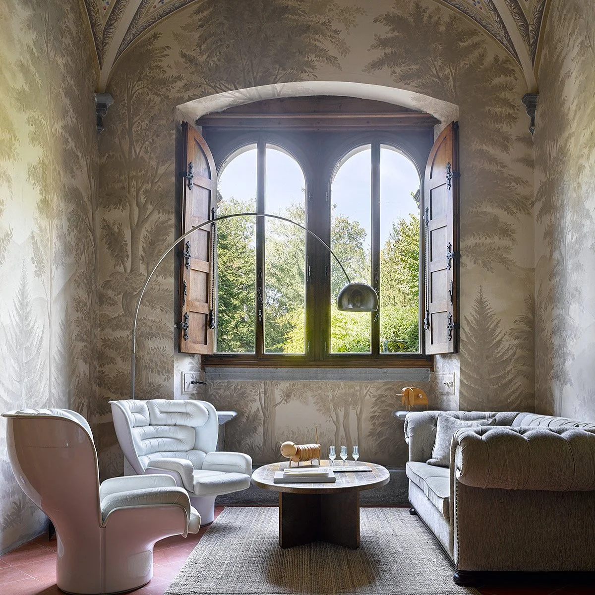 Interno di una stanza con pareti decorate a tema naturale, finestra grande con ante di legno aperte che mostra alberi verdi fuori, divano grigio, due poltrone bianche, tavolino rotondo con oggetti decorativi e bicchieri, lampada laterale sopra il div