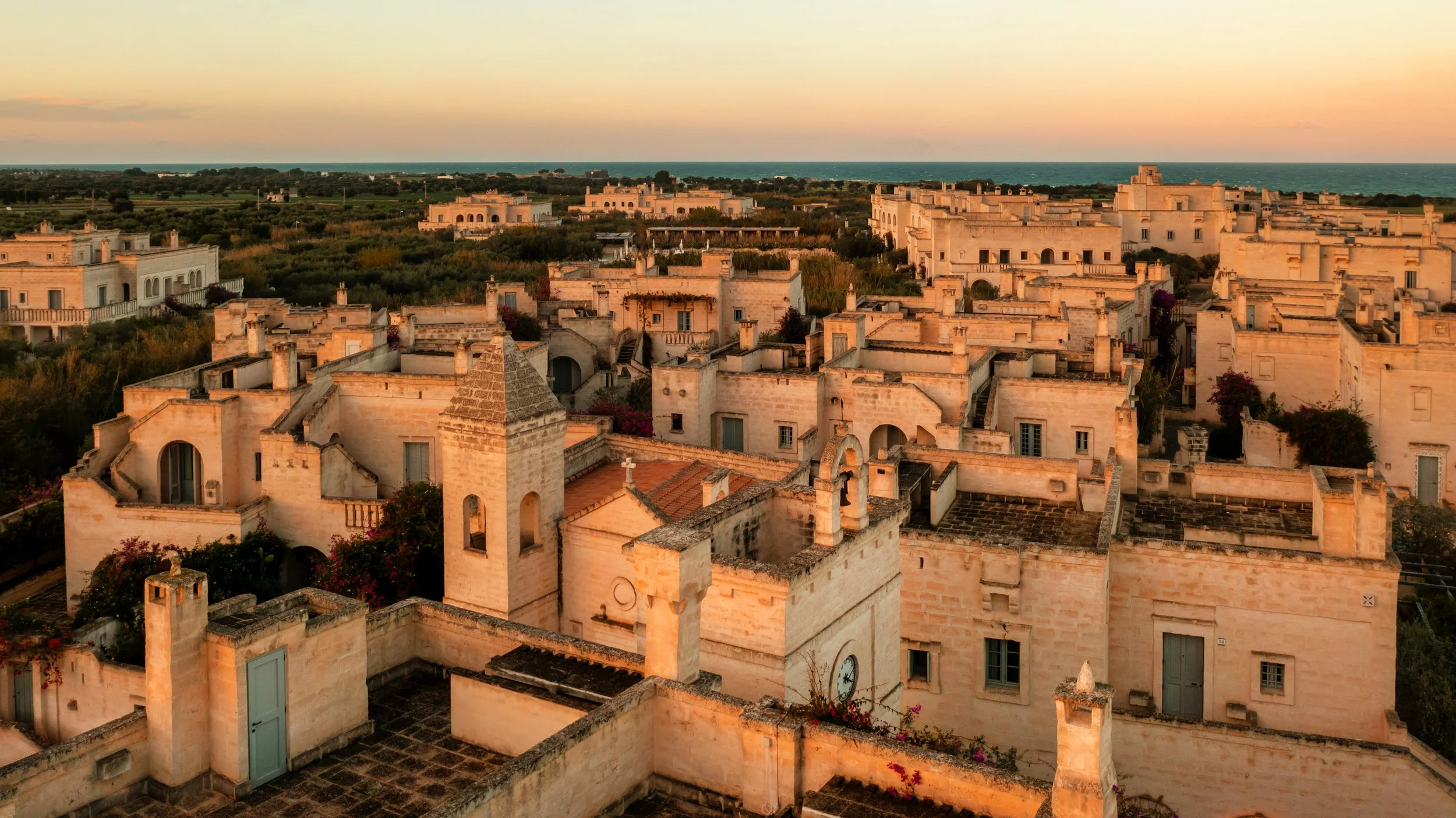 Veduta di un villaggio di case in pietra con tetti piatti e torri, al tramonto, sullo sfondo il mare e il cielo colorato di arancione e azzurro.