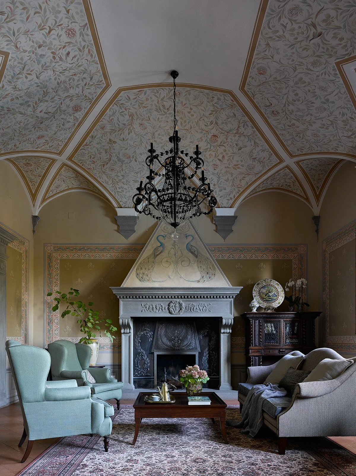 Salotto elegante con camino decorato, lampadario nero, divano grigio, poltrone azzurre, tavolino con vaso di fiori, arredamento vintage e pareti affrescate.