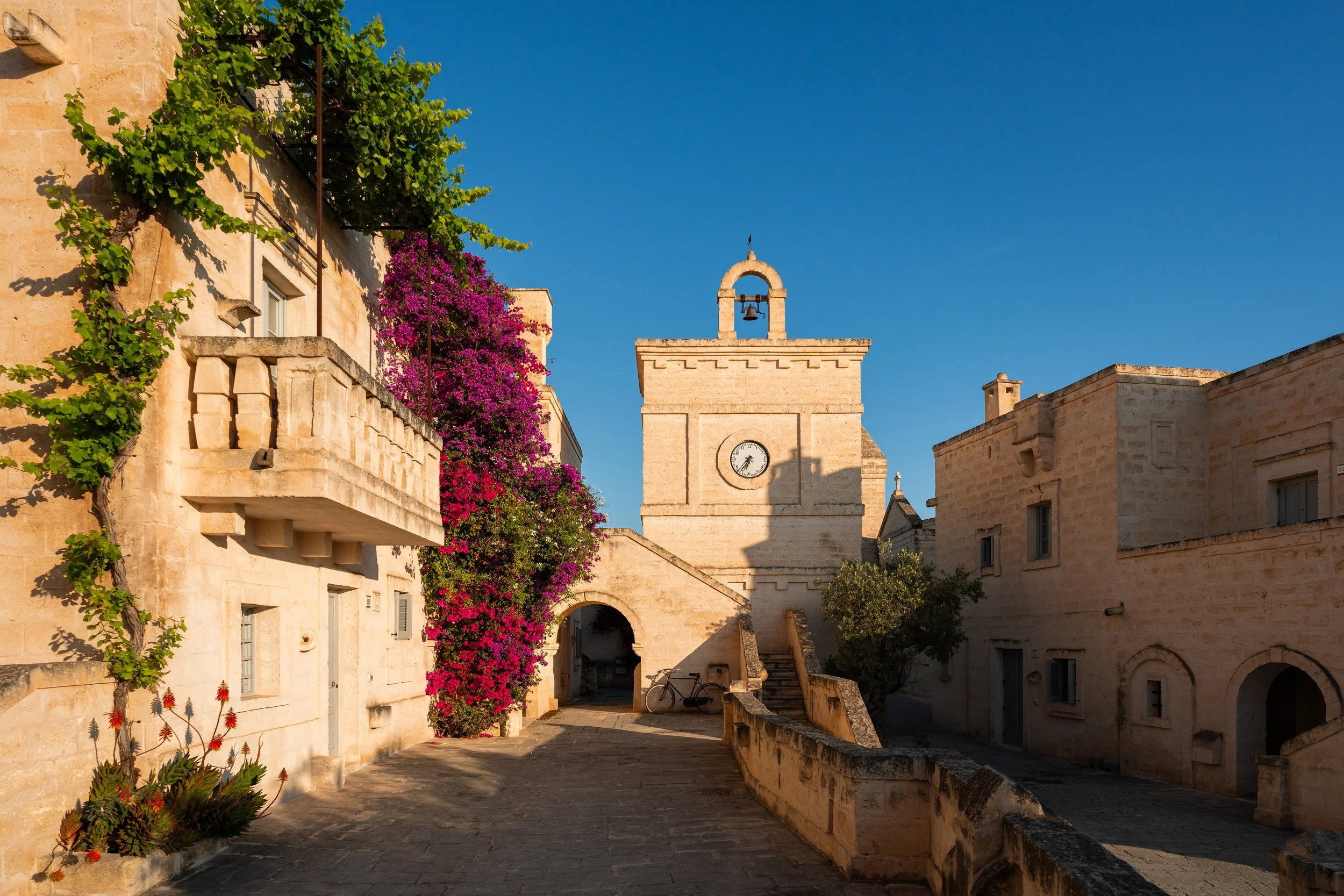 Vicolo con edifici in pietra chiara e fiori violetto-rosa, torre con orologio e campanile, in una giornata di sole con cielo blu.