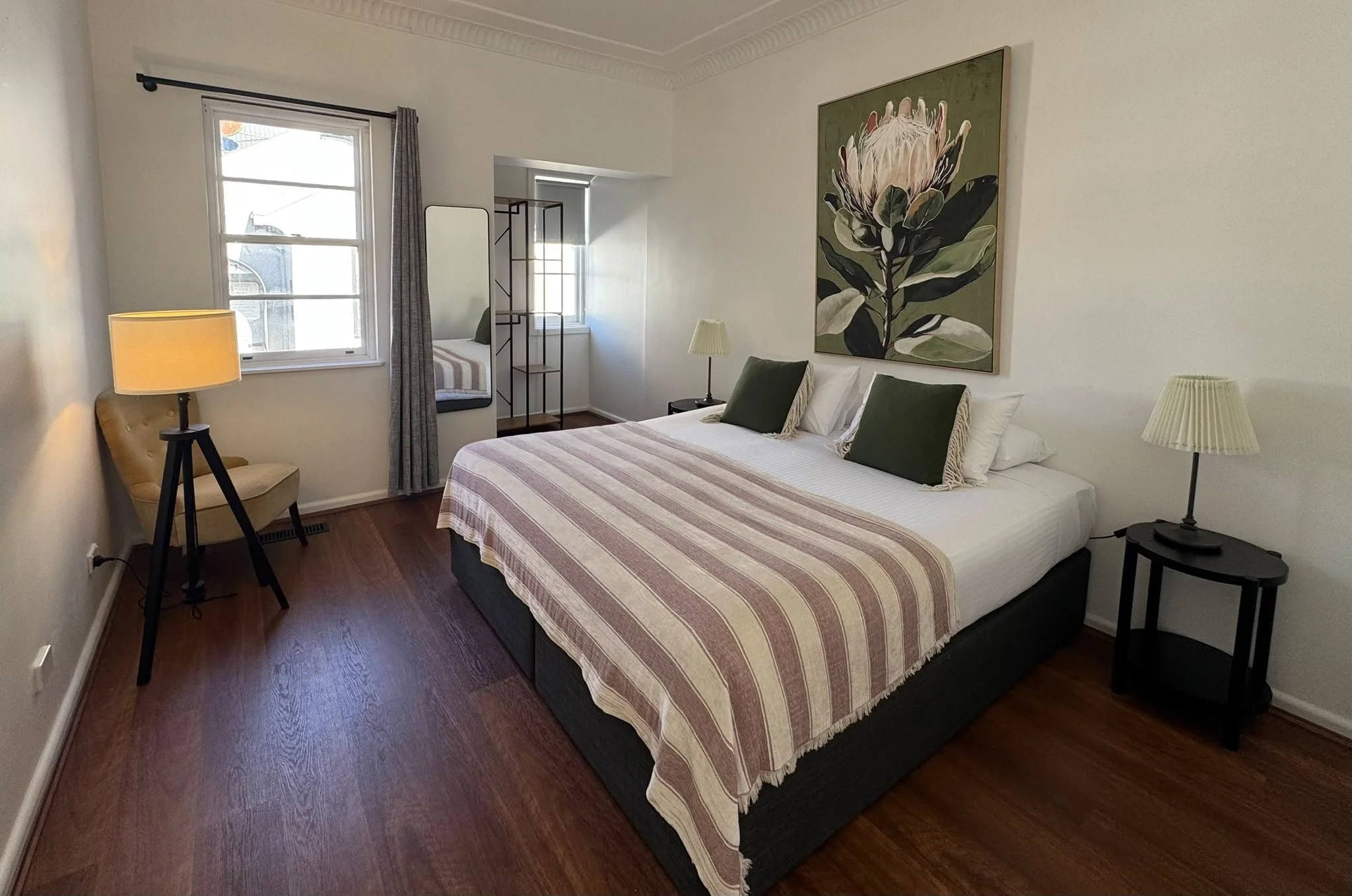 A bedroom with a large bed covered in a pink and white striped bedspread, two green pillows, and two white pillows. There are two nightstands with lamps on either side of the bed. A large floral painting is hung above the bed. A window with gray curtains is on the left, with a small room or alcove featuring a chair with striped upholstery and a small shelf or clothing rack nearby. The room has dark hardwood floors and white walls.