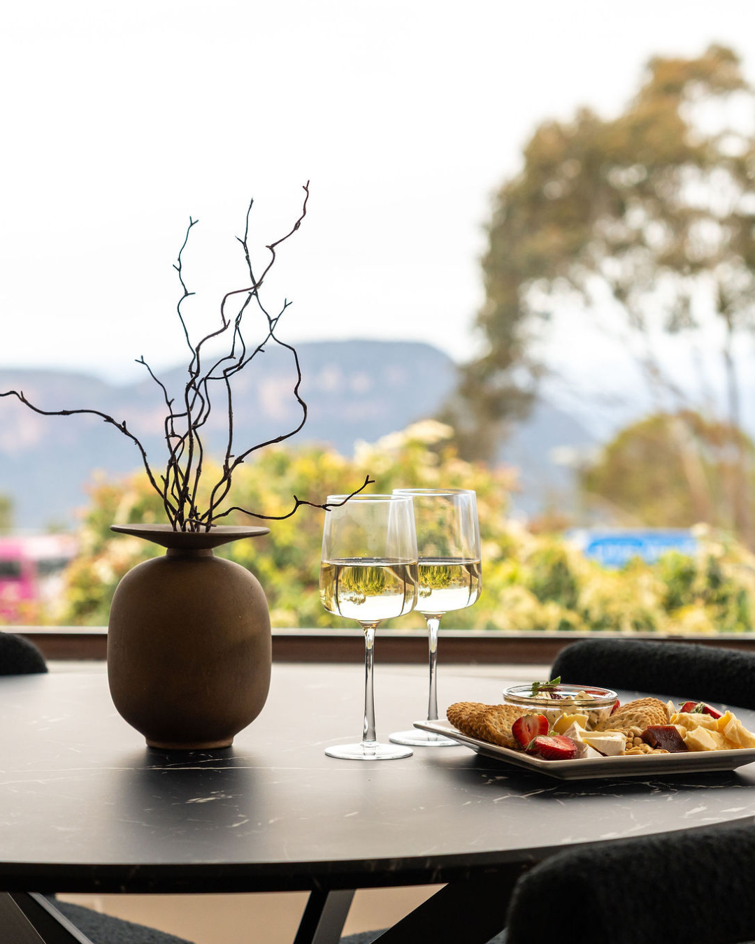 A table set with a decorative vase with twigs, two glasses of white wine, and a plate of cheese, crackers, strawberries, and other snacks, with a window showing trees and mountains outside.