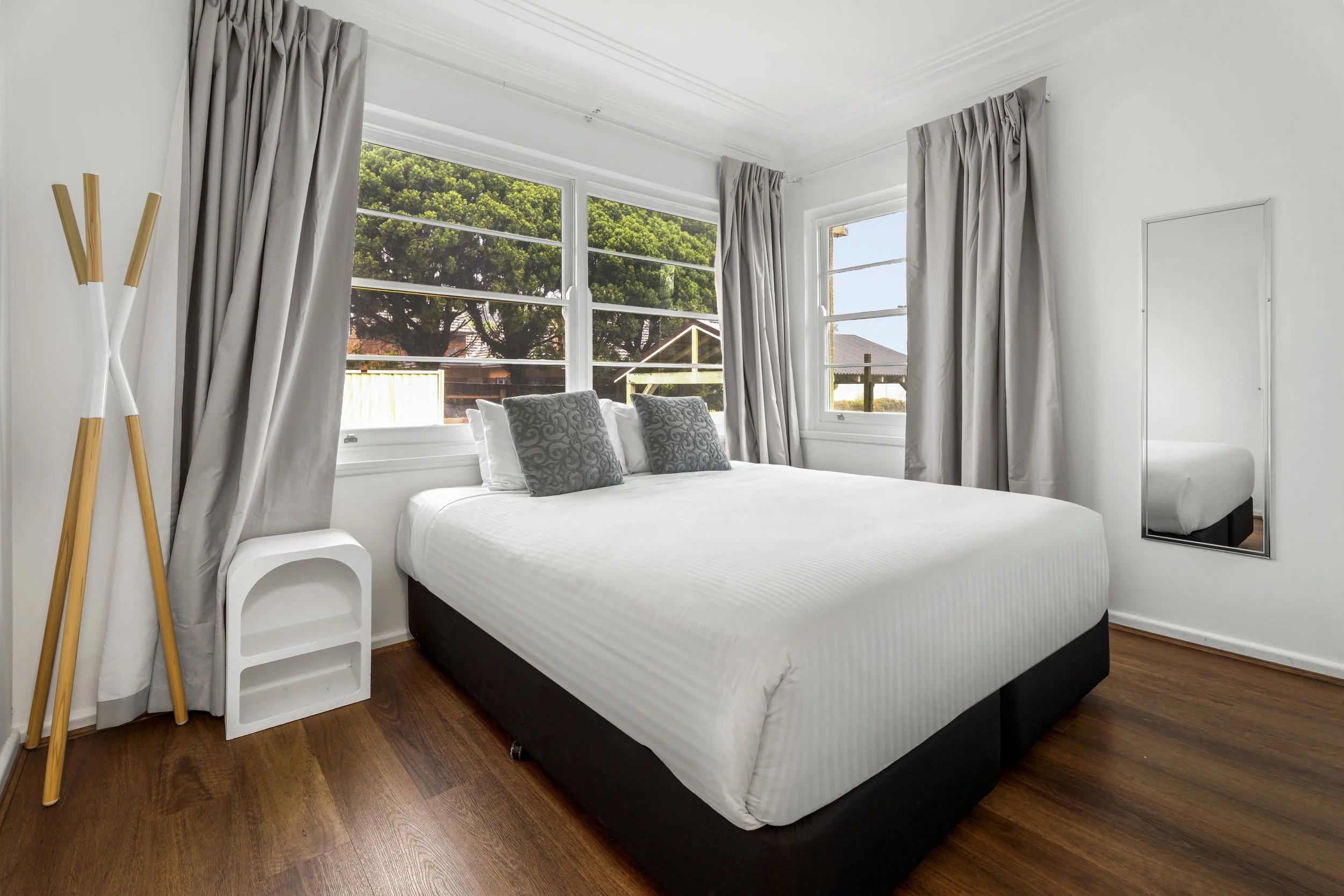 A modern bedroom with a white bed, two patterned pillows, large windows with gray curtains, hardwood floors, a white ladder shelf, and a mirror leaning against the wall.
