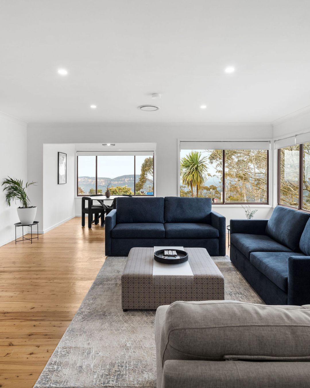 Living room with blue sofas, beige and gray area rug, wooden floor, white walls, large windows showing a view of trees and mountains, small side table with plant, and a dining area with a black table and chairs in the background.