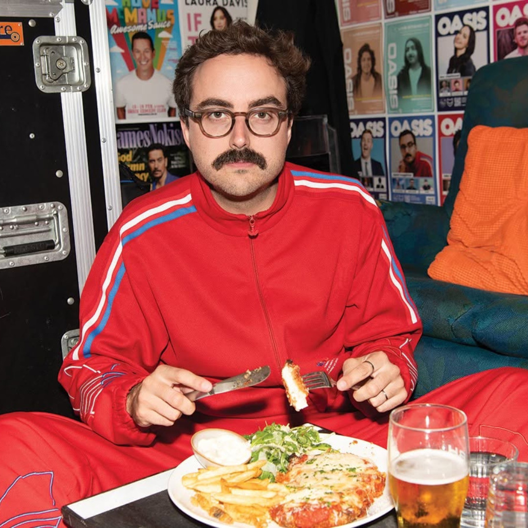 A man with glasses and a mustache wearing a red tracksuit with blue and white stripes, sitting on the floor in front of a table with food and drink, holding a fork and knife. The background features posters of actors and performers on the wall.