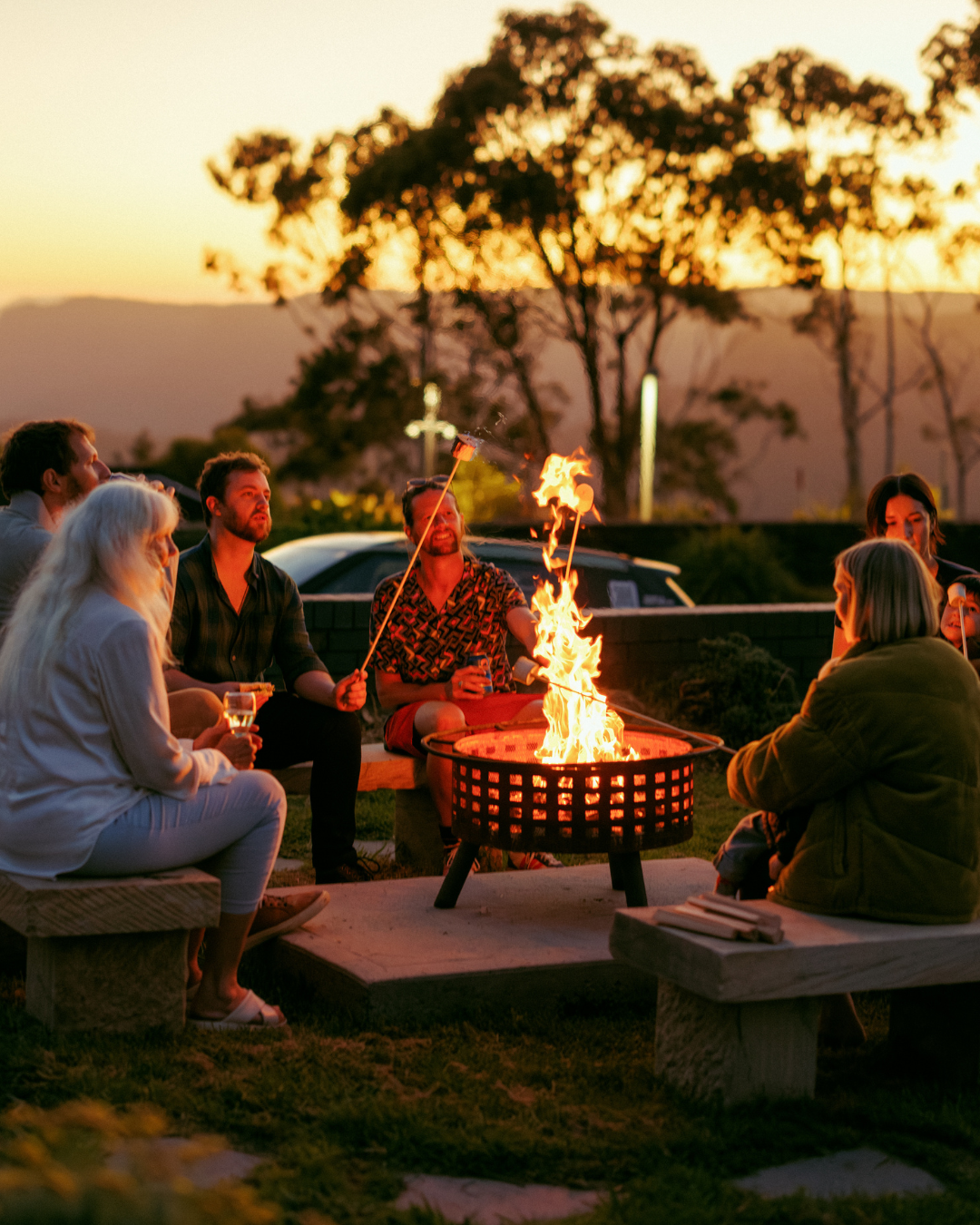 Group of people sitting around a fire pit outdoors during sunset, some roasting marshmallows on sticks.