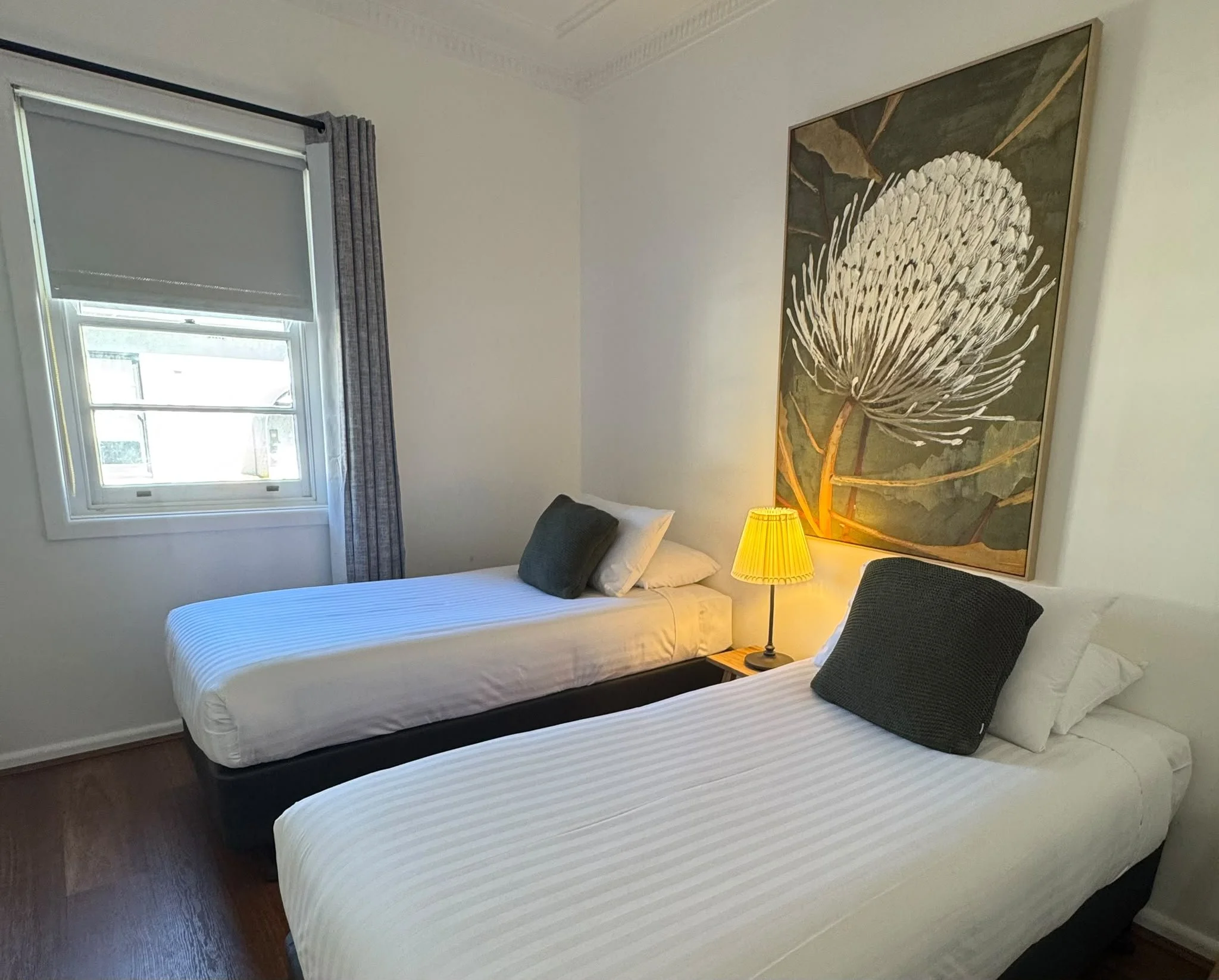 A bedroom with two single beds covered with white striped bedspreads, each with one dark pillow, a window with curtains, a large botanical artwork on the wall, a yellow bedside lamp, and a dark upholstered chair.