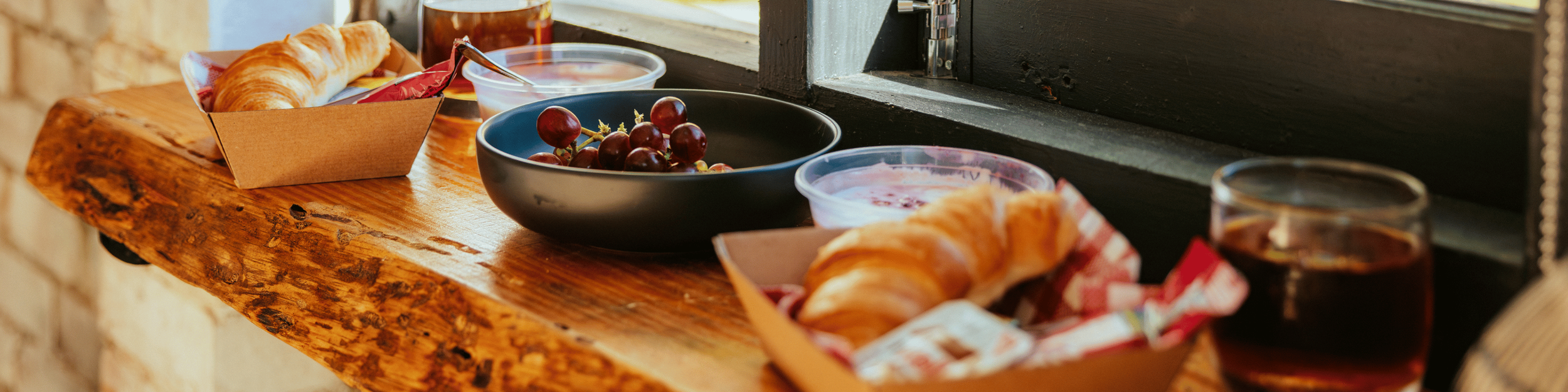 Fresh croissants in a cardboard tray, a bowl of red grapes, bowls of yogurt or sour cream, and glasses of dark-colored juice on a rustic wooden table near a window.