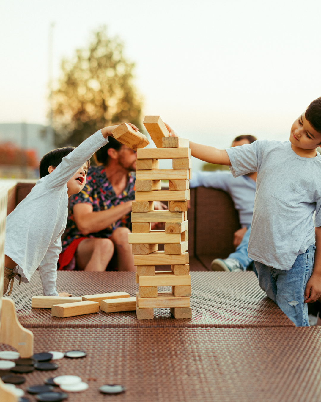 Kids playing Jenga outdoors at sunset with adults sitting in the background.