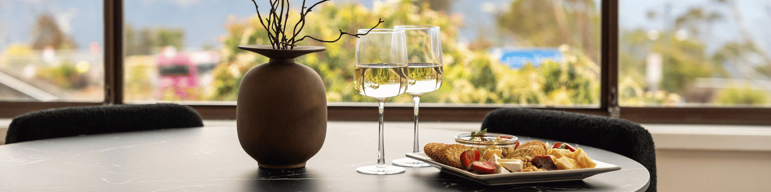 A table with two glasses of white wine, a plate with cheese, crackers, and strawberries, and a brown vase with decorative branches, set near a window with a blurred outdoor view.