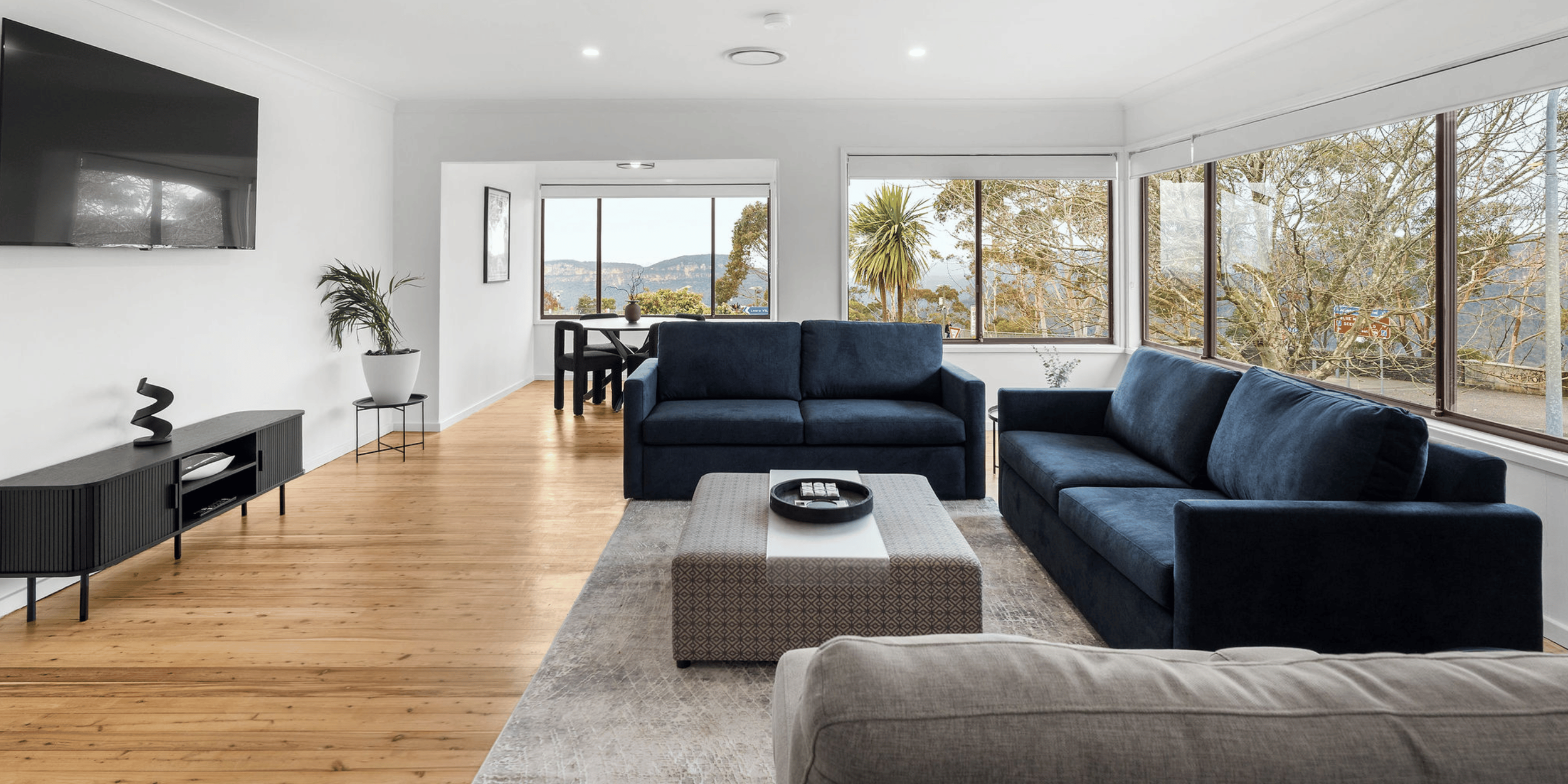 A bright living room with wooden flooring, white walls, large windows showing trees and mountains outside, and modern dark blue sofas. There is a light-colored ottoman, a black media console with decorative objects, and a small dining area with black