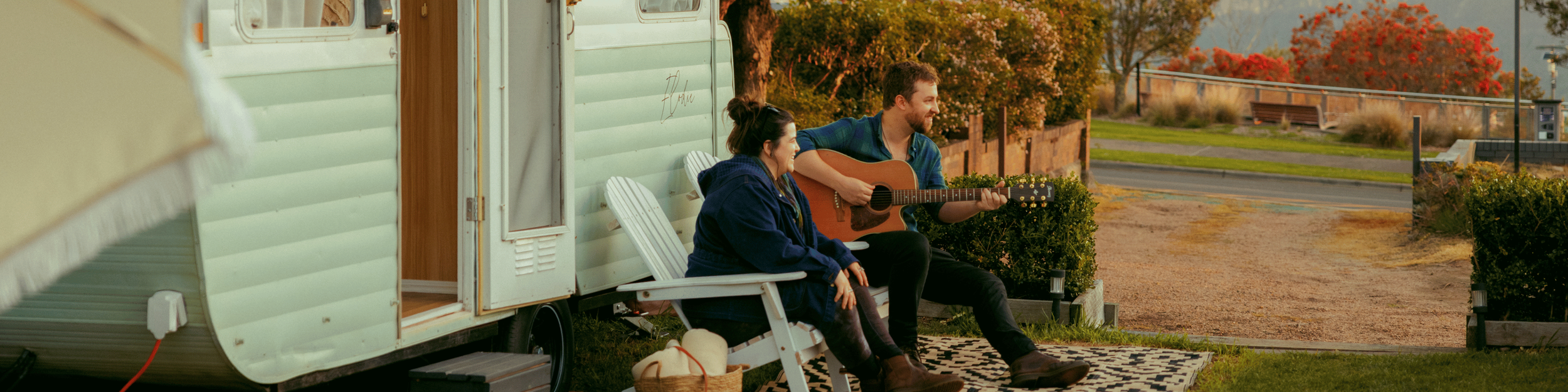 A man and woman sitting outside a vintage camper trailer; the man is playing a guitar, and the woman is listening, with a scenic background of trees and mountains.