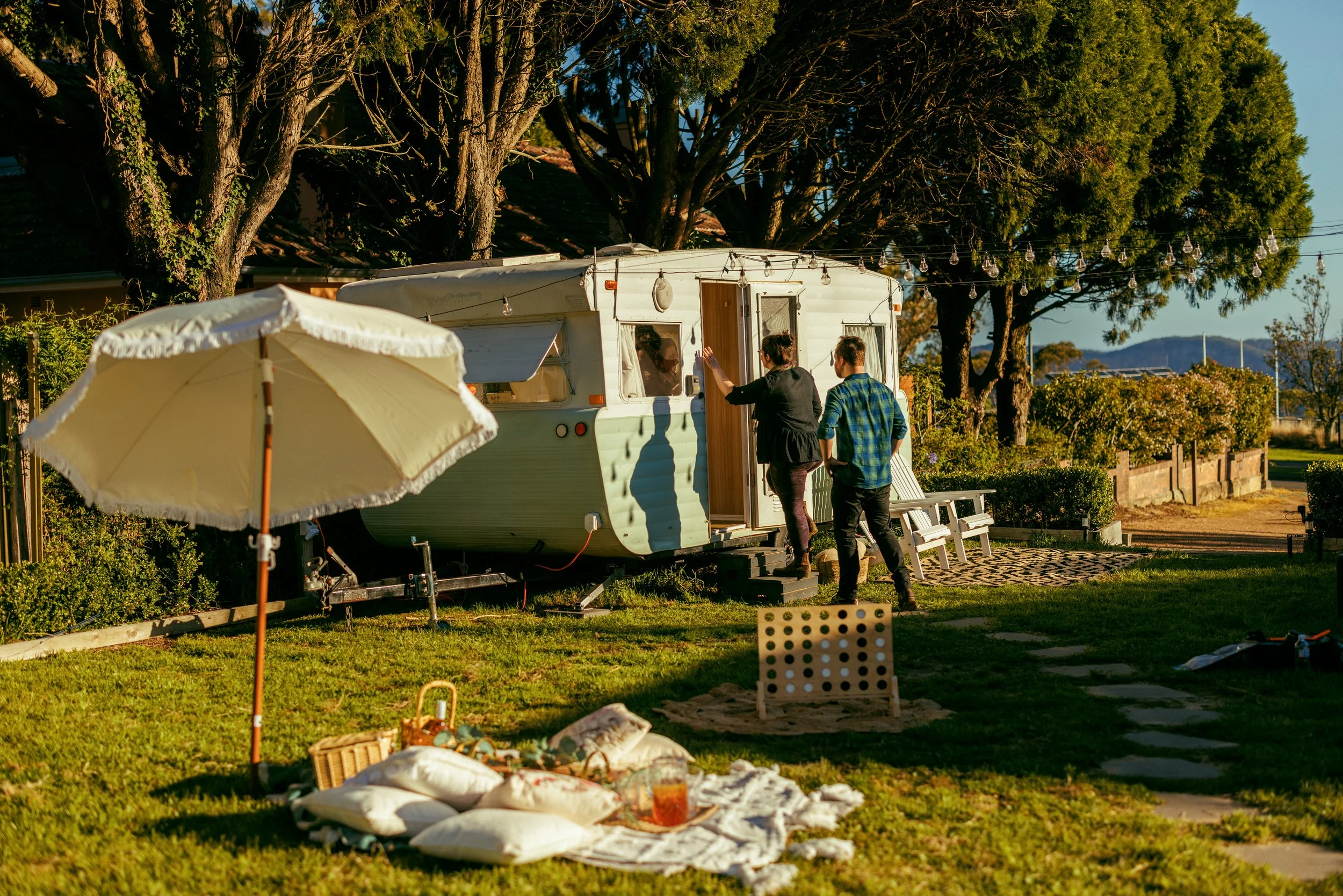 A vintage camper trailer park with two people standing outside, one gesturing. An umbrella, pillows, and blankets are laid out on the grass, and string lights are hung above. Tall trees surround the scene on a sunny day.